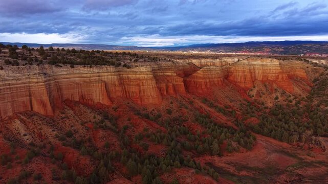 Dramatic red sandstone canyon cliffs under a moody overcast sky at dusk, with scattered juniper trees, layered rock formations, and a distant valley town in the American Southwest