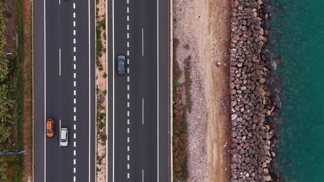 Overhead view of a multi-lane coastal road with a single white car driving alongside a rocky breakwater and turquoise sea, separated by a sandy strip