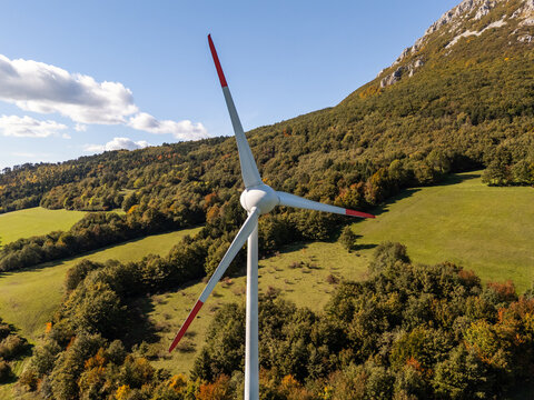 A wind turbine stands tall in a green field surrounded by hills and trees. The scene shows a clear sky and natural surroundings.