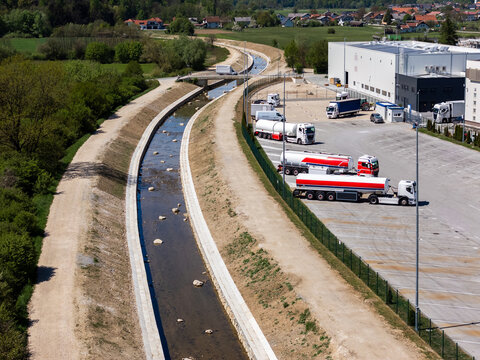 River restoration work is visible with channel cleaning and widening near a warehouse. Trucks are parked nearby in the industrial area.