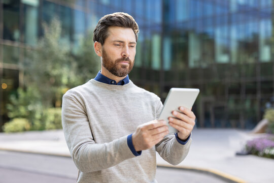 A serious young man is standing on the street and looking at the tablet he is holding