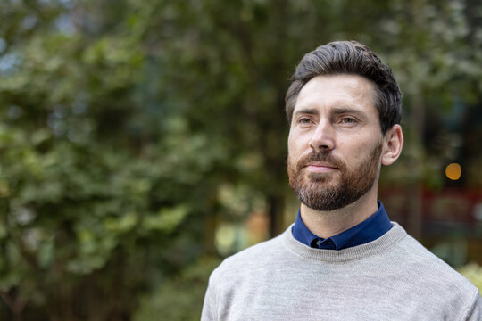 A close-up photo of a young man standing in a park with his right profile and looking seriously ahead