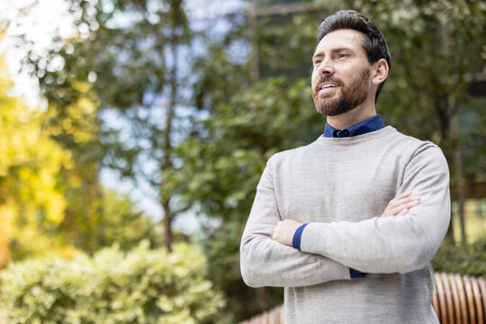 Close-up photo of a young confident businessman outside, standing with his arms crossed on his chest and looking into the distance