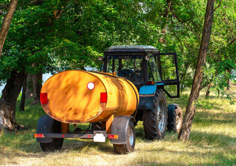Rear view of a blue farm tractor attached to a yellow water tank trailer, standing on dry grass in a shaded wooded rural setting © ravital