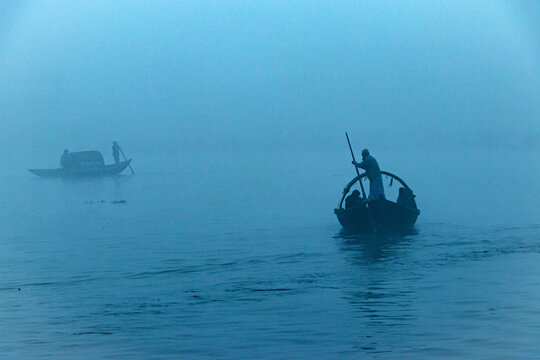 Narayanganj, Dhaka, Bangladesh - 24 January 2021: View of traditional wooden boats crossing a river in dense winter fog with people rowing.