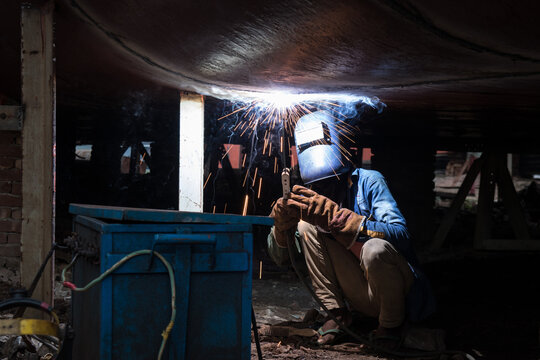 Dhaka, Bangladesh - 04 November 2018: View of a worker in a welding mask welding a ship hull in a shipyard along the Buriganga River, with bright sparks flying in the dark workspace.