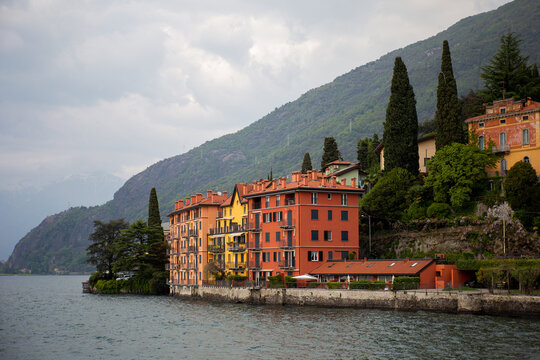 Vivid red and yellow residential buildings situated right on the water edge of Bellano with cypress trees and misty mountain slopes in the background on Lake Como.