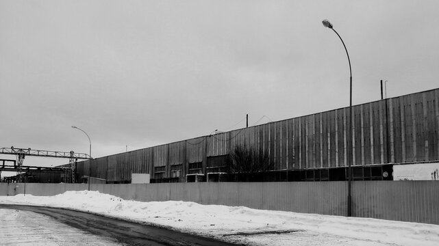 Black and white industrial landscape with a long corrugated metal fence and factory building under a cloudy sky