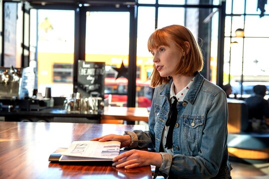 Person with red hair in a cafe, holding a magazine at a table.