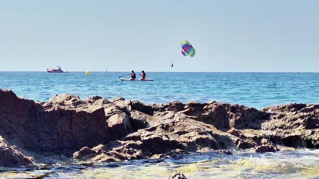 August 2026. Lloret de Mar, Spain. Parasailing activity and people paddleboarding near a rocky coastline on a sunny summer day