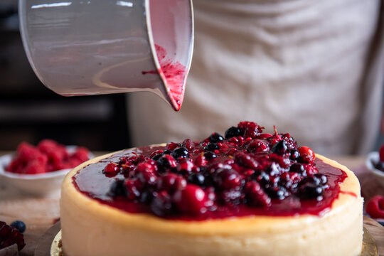 Berry sauce being poured over a cheesecake in a kitchen setting.