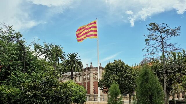 August 2026. Barcelona, Spain. Catalan flag waving on a tall flagpole above historic architecture and green trees on a sunny summer day