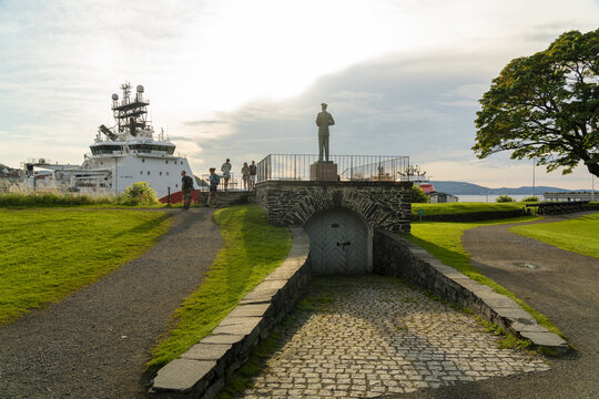 Bergen, Norway - 17 July 2025: View of the statue of King Haakon VII at Bergenhus Fortress with a large ship in the harbor and people walking on the grounds during the golden hour.