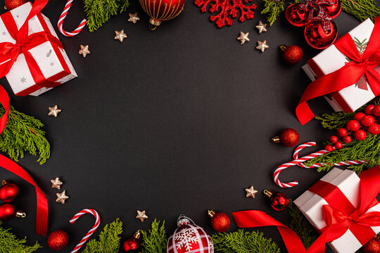 A festive Christmas themed flat lay image with red and white gift boxes, baubles, candy canes, glittering stars, red ribbon, and green fir branches forming a decorative border on a black background.