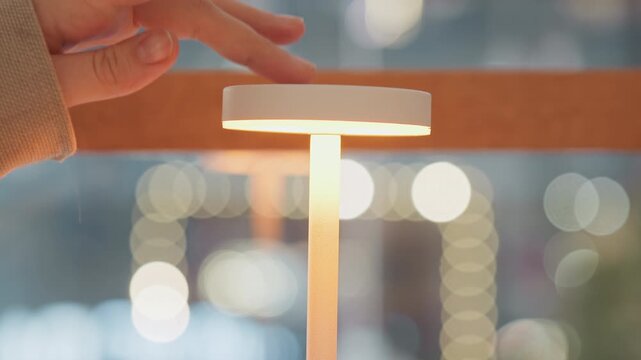 Close up of hand interacting with and turning on elegant white LED lamp emitting soft light in modern indoor setting. scene features warm ambient lighting effects from blurred background