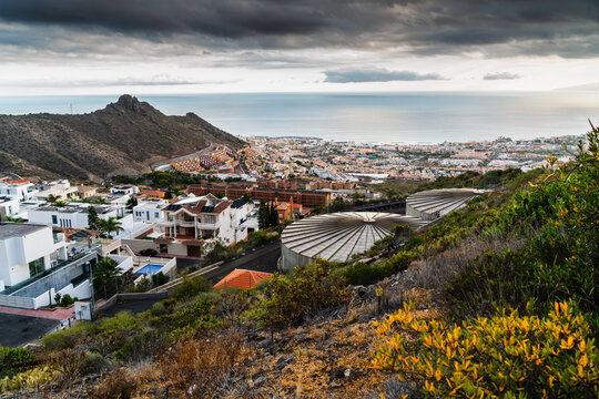 Costa Adeje, Canary Islands, Spain - 26 June 2026: View of the coastal town of Costa Adeje with white villas, large circular water tanks, and the Atlantic Ocean under a cloudy sky.