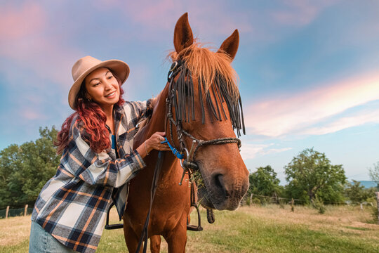 Against a dramatic pastel sunset sky, a smiling woman strokes her chestnut horse at a hilltop rural pasture. Equestrian lifestyle concept.