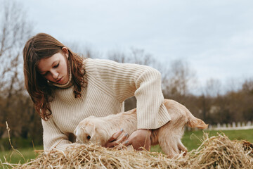 Naklejka premium A woman in a cream knit sweater bends beside hay, gently guiding a young goat kid in a sunny outdoor farm scene. A candid lifestyle moment of animal care and bonding.
