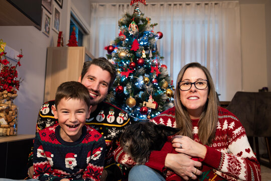 Happy family wearing festive ugly christmas sweaters and jeans, sitting together on the floor with their dog in front of a decorated christmas tree with lights and ornaments