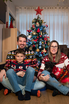 Happy family &mdash; parents, young boy and small dog in festive sweaters &mdash; sitting on the living room floor by a lit, decorated christmas tree, smiling and cozy together