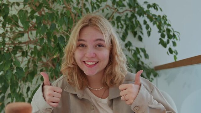 Close up of young woman with blonde hair wearing layered necklaces and beige jacket seated in front of green foliage. Woman maintains direct eye contact while giving two thumbs up gestures