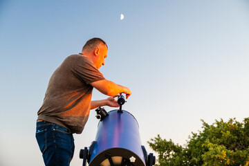 a man adjusting a large blue telescope outdoors against a twilight sky with a visible crescent moon. © Andrey