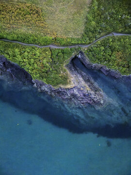 Aerial view of the rugged coastline at Hawker's Cove, featuring dark rock formations, turquoise sea, lush vegetation, and a coastal path in Padstow, Cornwall, United Kingdom.