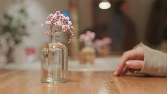 Close up of clear glass vase with pink flowers on wooden table. Hand in long sleeve reaches towards empty bottle then retracts soft lighting and blurred background with another similar