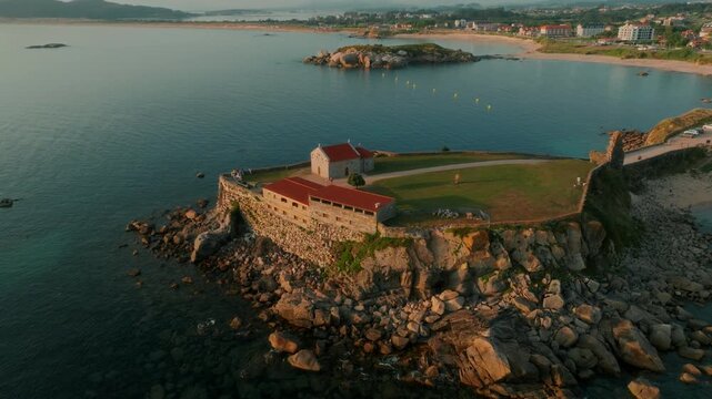 erial view of a historic stone hermitage perched on a rugged coastal promontory at golden hour, overlooking the serene blue waters of the bay and sandy shoreline