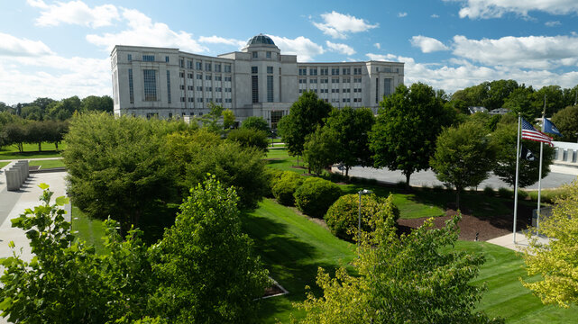 Aerial view of the Michigan Supreme Court building surrounded by lush green trees, manicured lawns, and American flags under a bright blue sky in Lansing, Michigan, United States.
