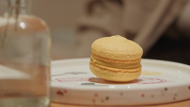 Close up of yellow macaron on white plate with floral patterns accompanied by glassware and soft lighting. scene features consistent indoor setting details including wooden table surface