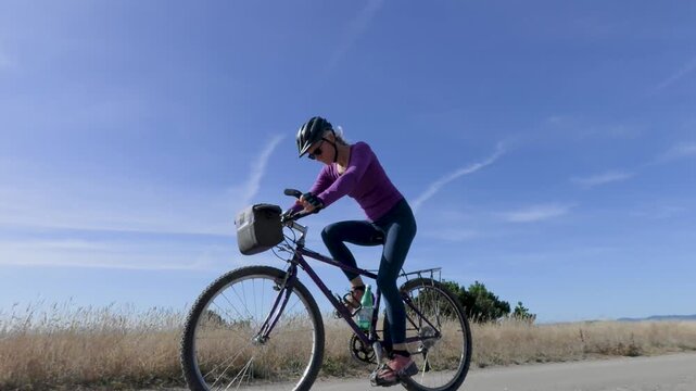 Mature woman rides a bike on a paved trail in Fort Bragg near the ocean enjoying the clear blue sky and nature around her.