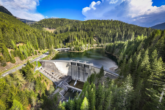 Aerial view of Sperre Paal Bodendorf dam surrounded by dense coniferous forests, a winding road, and a calm reservoir under a bright blue sky in Murau, Styria, Austria.