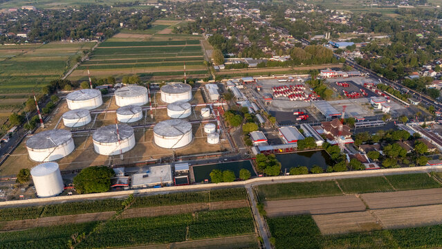 Aerial view of an industrial fuel storage facility with large white cylindrical tanks and a fleet of red tanker trucks parked in a lot, surrounded by agricultural fields in Indonesia.