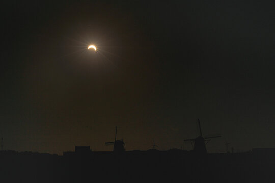 Partial solar eclipse over silhouetted windmills at dusk with dark evening sky