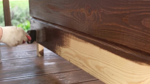 Hand in glove painting wooden box with brown oil stain brush. Worker applying protective varnish on pine timber planks surface, DIY woodwork. Wood staining process. Backyard Woodworking, Maintenance.