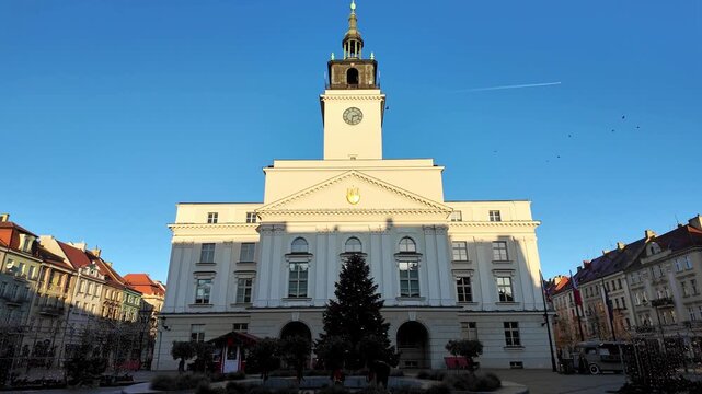Video of neoclassical town hall in Kalisz with tall clock tower, filmed from main market square featuring Christmas tree and surrounding historic tenement houses under clear blue sky in daylight.