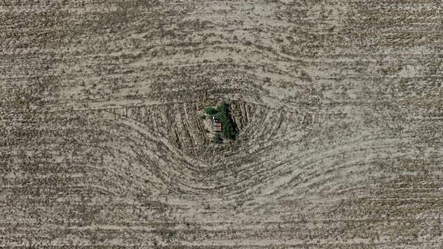 Aerial view of a small shed surrounded by green trees in the center of a large, dry, plowed agricultural field with concentric patterns in Frederick, Colorado, United States.