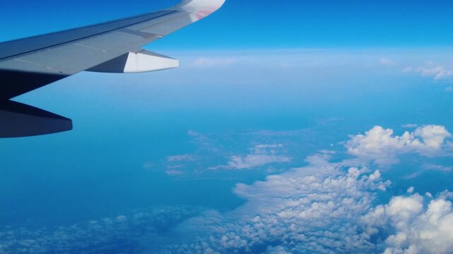 airplane wing extends over open blue sea distant coastline emerges through scattered clouds window seat view mid flight afternoon light calm horizon wingtip visible sunlit wing surface reflects light