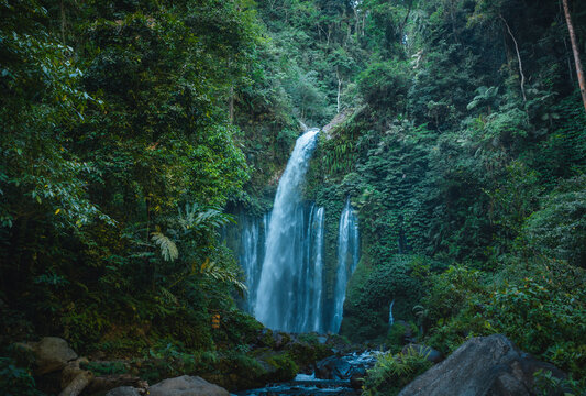 Aerial view of Tiu Kelep Waterfall cascading down into a rocky pool surrounded by lush tropical rainforest and dense jungle vegetation in Bayan, West Nusa Tenggara, Indonesia.