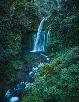 Aerial view of Tiu Kelep Waterfall cascading into a rocky riverbed surrounded by lush tropical rainforest in Bayan, West Nusa Tenggara, Indonesia.