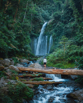Aerial view of Tiu Kelep Waterfall cascading into a rocky river with a person standing on a fallen log in the lush tropical rainforest, Bayan, West Nusa Tenggara, Indonesia.