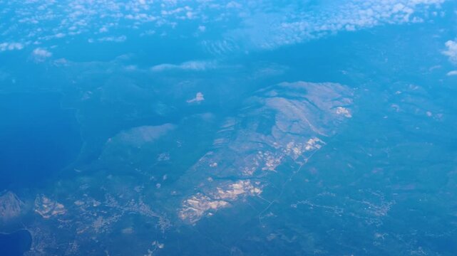 rugged coastal terrain unfolds far below patchwork farmland climbs hillsides scattered clouds drift overhead window view from airplane cruise altitude sunlit landscape passenger perspective