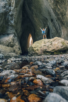 Woman standing on a rock inside a huge cave, arms open, celebrating successful exploration and adventure in nature