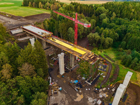 Aerial view of the Lezky bridge construction site, featuring concrete pillars, a yellow bridge deck, and a red tower crane amidst forest and fields in Lubenec, Usti nad Labem Region, Czechia.