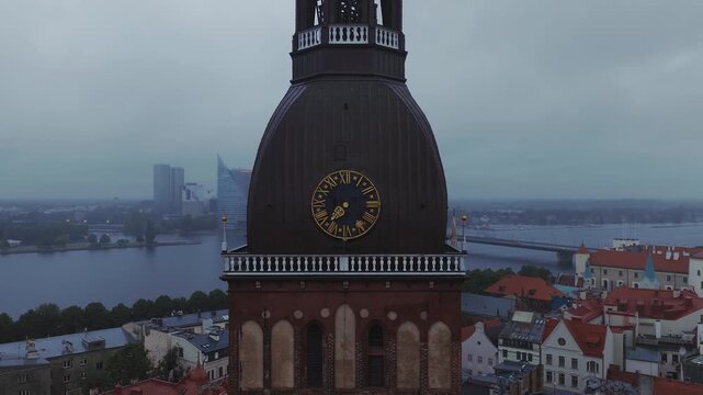 An aerial shot circling the historic Riga Cathedral clock tower with views over the Daugava River and cityscape under an overcast sky in Latvia.