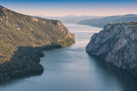 Calm blue Danube river stretches deep into a forested limestone gorge of Iron Gates framed by sheer rocky cliffs and distant hazy ridgelines. Scenic destination in Serbia and Romania concept.
