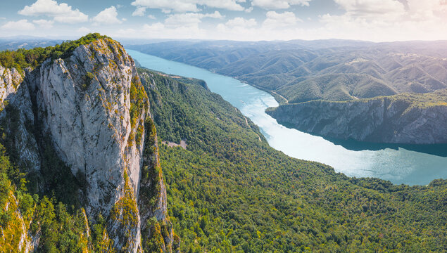 Sheer vertical limestone pinnacle rises dramatically beside a turquoise Danube river winding through a vast green canyon of Iron Gates. Adventure concept.