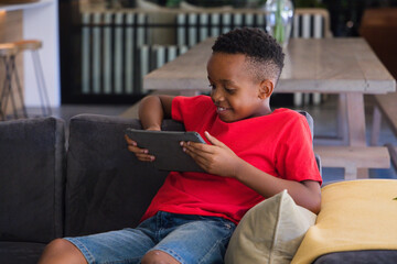 African American child sitting on gray sofa at home holding tablet wearing red tee denim shorts © WavebreakMediaMicro