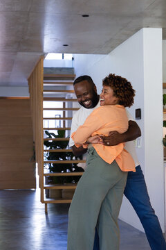 African American couple wearing casual clothes embracing and laughing at home near wooden staircase